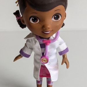 May include: A Doc McStuffins figurine wearing a white lab coat, a pink stethoscope, and pink shoes. The doll has brown skin, dark hair, and a pink headband. The figurine is a collectible item.