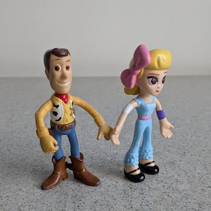 May include: Toy Story characters Woody and a female character with a pink bow in her hair. Woody is wearing his signature brown cowboy hat, vest, and jeans. The female character is wearing a light blue jumpsuit with a purple band on her arm.