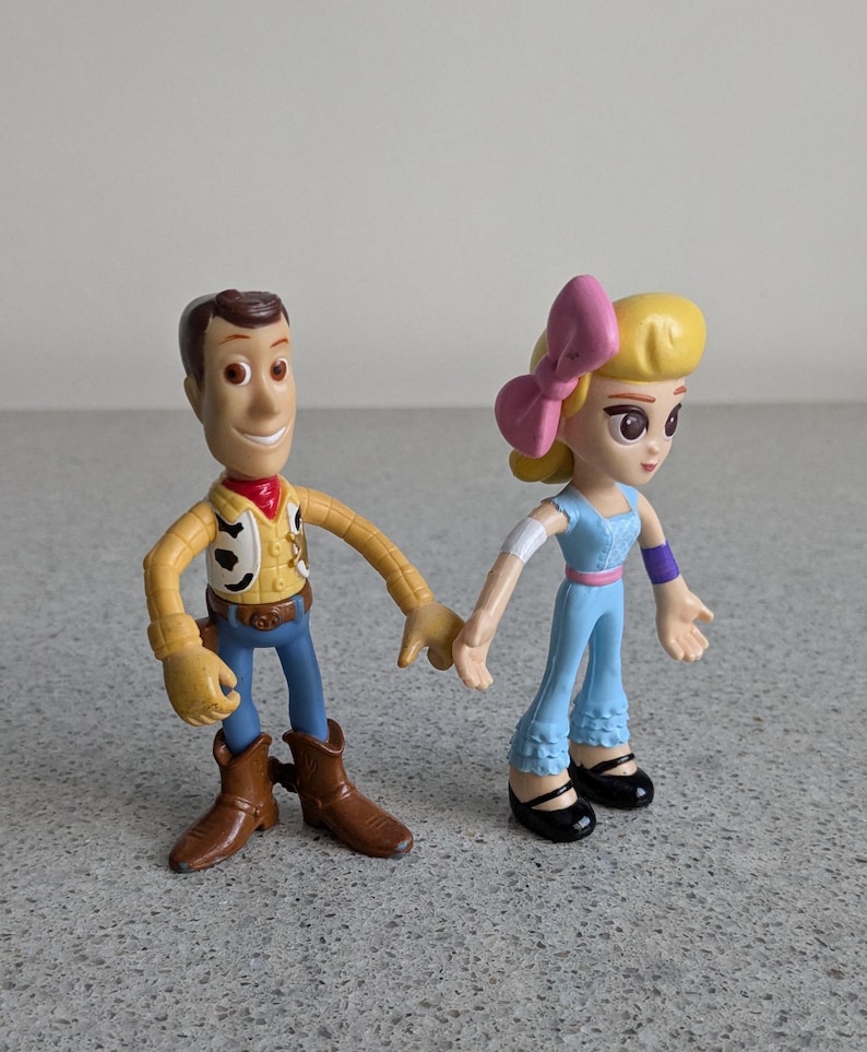 May include: Toy Story characters Woody and a female character with a pink bow in her hair. Woody is wearing his signature brown cowboy hat, vest, and jeans. The female character is wearing a light blue jumpsuit with a purple band on her arm.