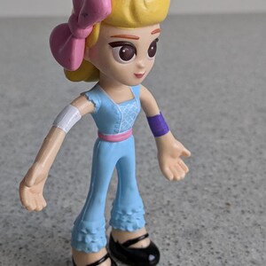 May include: A small plastic doll with blonde hair, a pink bow, and a blue dress. The doll has brown eyes and a light skin tone. The doll is wearing black shoes.