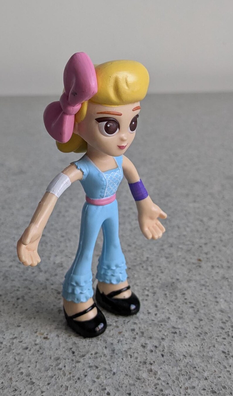 May include: A small plastic doll with blonde hair, a pink bow, and a blue dress. The doll has brown eyes and a light skin tone. The doll is wearing black shoes.