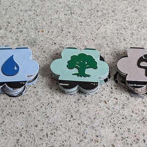 May include: A collection of five flower-shaped game tokens. Each token features a different color and symbol: red flame, blue water drop, green tree, gray skull, and yellow sun. The tokens are stacked and appear to be made of cardboard.
