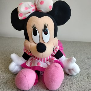 Puede incluir: Un vestido de lunares rosa y blanco con un lazo rosa en la cabeza. El juguete de peluche est&aacute; sentado con los brazos extendidos. El juguete tiene orejas negras y una nariz negra.