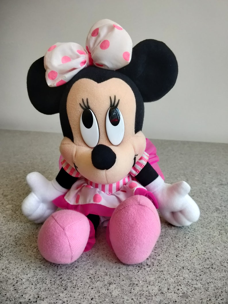 Puede incluir: Un vestido de lunares rosa y blanco con un lazo rosa en la cabeza. El juguete de peluche est&aacute; sentado con los brazos extendidos. El juguete tiene orejas negras y una nariz negra.