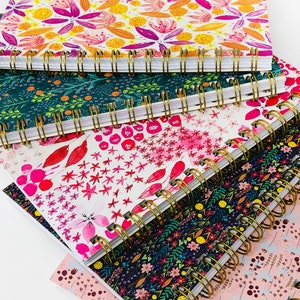 May include: A stack of five spiral bound notebooks with colorful floral patterns. The covers feature a variety of flowers, leaves, and abstract designs in shades of pink, green, yellow, and blue.