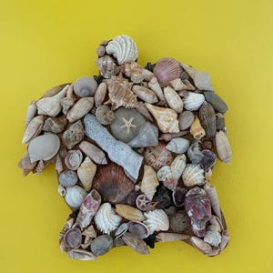 Puede incluir: Una colección de diversas conchas marinas, incluyendo caracolas y estrellas de mar, dispuestas en forma de persona sobre un fondo amarillo. Las conchas son en tonos de blanco, beige y marrón, creando una estética texturizada y natural. La obra es una pieza decorativa única.