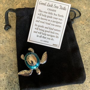 Puede incluir: Un dije de tortuga marina plateado con un caparazón turquesa y plateado descansa sobre una bolsa de terciopelo negro. El dije va acompañado de una tarjeta que dice "Good Luck Sea Turtle Charm". El dije de tortuga es un accesorio decorativo.
