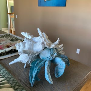 Op de afbeelding: Een decoratieve sculptuur van een heremietkreeft. De krab heeft een lichtblauw lichaam en grote klauwen, met een witte schelp. De sculptuur staat op een houten tafel. Op de achtergrond een schilderij van een zeeschildpad.