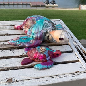 May include: Two sea turtle figurines crafted from resin with a swirling multi-colored pattern of pink, purple, and teal. The larger turtle features a clear head and shell with embedded shell accents, while the smaller turtle has a solid shell.