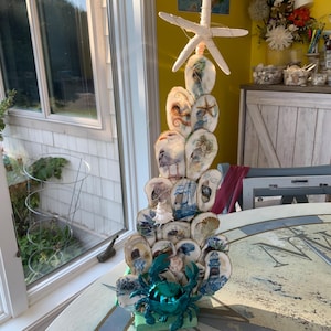 May include: A decorative coastal-themed sculpture resembling a tree, crafted from seashells and topped with a starfish. The seashells are adorned with nautical imagery and a turquoise crab sits at the base. The sculpture is displayed on a round table.