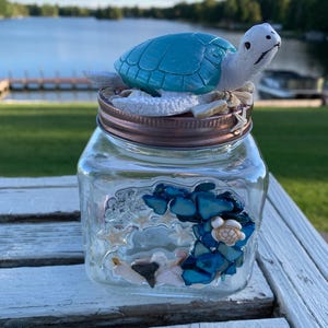 May include: A clear glass jar with a copper-coloured lid, decorated with blue sea glass, seashells, and a small turtle figurine with a turquoise shell. The jar is sitting on a white wooden surface.