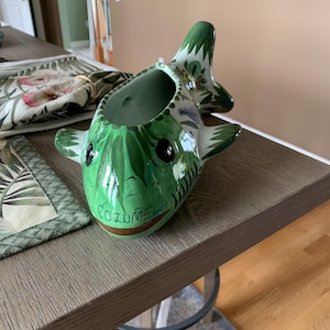 May include: A green and white ceramic fish-shaped planter with the word "COZUMEL" painted on the front. The planter has black eyes and is decorated with leaf and floral patterns. The planter is sitting on a wooden table.