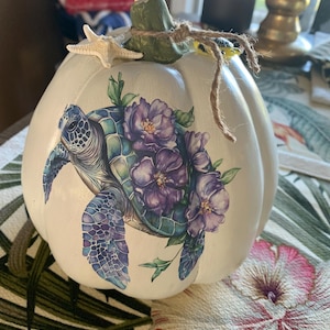 May include: A white decorative pumpkin featuring a detailed illustration of a sea turtle surrounded by purple flowers. The pumpkin has a green stem tied with a piece of twine and is accented with a small white starfish and a tiny turtle figure.