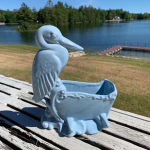 May include: A light blue ceramic planter featuring a sculpted heron standing on a base next to an open, rectangular basin with decorative embossed scrollwork along the top rim and four small feet at the bottom of the structure.