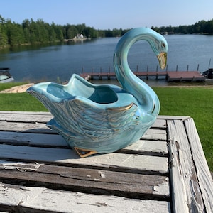 Könnte beinhalten: Eine hellblaue Schwanenfigur aus Keramik mit irisierender Oberfläche und goldfarbenen Akzenten an Schnabel, Flügelgefieder und Sockel. Der Schwan hat einen geschwungenen Hals und eine offene, hohle Mitte als dekoratives Gefäß.