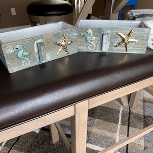 May include: Two rectangular wooden boxes with a weathered gray finish. Each box features a decorative seahorse adorned with blue and green rhinestones and a shiny gold-toned starfish emblem attached to the front exterior side panels.