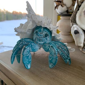 May include: A teal crab sculpture with a textured surface and white accents, emerging from a large, textured white shell. The crab has detailed claws and a detailed face. The sculpture is displayed on a wooden surface.