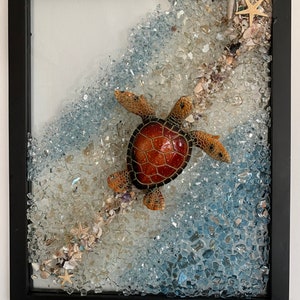 May include: A framed artwork featuring a sea turtle figurine swimming through a wave of blue and white glass mosaic tiles. The mosaic is accented with seashells and starfish.