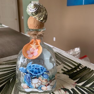 May include: A clear glass bottle with a cork stopper topped by a green and white spiral seashell. The bottle features a graphic of a blue sea turtle, seashells, and a hanging orange shell with a starfish and pink flower print tied with twine.