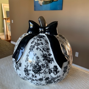 May include: A ceramic pumpkin-shaped decorative object featuring a white base with a black floral toile pattern. The pumpkin is topped with a glossy black bow and a metallic stem, creating a high-contrast look with intricate botanical details.