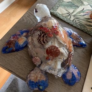 Puede incluir: Figura decorativa de tortuga hecha de resina transparente, rellena de conchas marinas y adornada con una mariposa roja enjoyada. Las aletas y la cabeza presentan detalles en azul y blanco. Un artículo de decoración del hogar único.