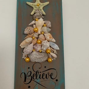 May include: A rectangular wooden plaque with a teal and brown painted background. A Christmas tree is crafted from various seashells and gold-colored beads. A starfish tops the tree. The word "Believe" is written in cursive.