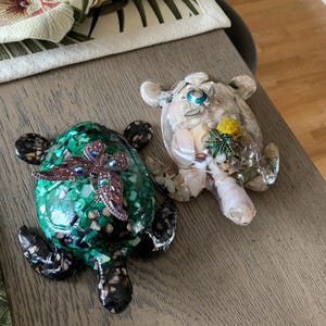 May include: Two decorative turtle ornaments. One is green with a jewelled dragonfly, the other is clear and filled with shells and a yellow flower. Both have detailed flippers and heads.