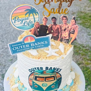 May include: A white frosted cake decorated with Outer Banks themed images and text. The cake has a gold topper that reads "Happy 12th Birthday Sadie". The cake is decorated with edible sand, seashells, and images of the Outer Banks cast and logo.