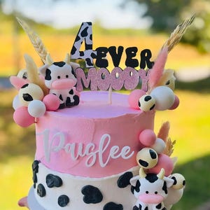 May include: A two-tiered pink cake decorated with cow-themed accents. The cake features a pink frosting base, white frosting with black cow spots, and a topper that reads "4 Ever Moody". Additional decorations include cow figurines and pink and white spheres.