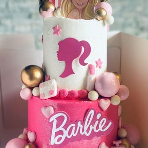 Op de afbeelding: Een tweelaagse taart met witte en felroze glazuur. De bovenste laag heeft een Barbie-silhouet en een topper van een blond personage met een tiara en regenboog. De onderste laag toont het Barbie-logo, een handtas en Happy 6th Birthday Brynlee.