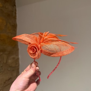 Puede incluir: Tocado naranja con detalles de rosas y plumas. El tocado presenta un diseño de malla y está sujeto a una diadema fina. El color es naranja vibrante, adecuado para ocasiones especiales.