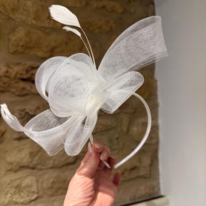 Op de afbeelding: Een witte haarband met een grote, getextureerde strik en delicate veermotieven. De strik is gemaakt van een transparante, netachtige stof en de haarband is een eenvoudige, dunne band. Dit elegante hoofddeksel is geschikt voor speciale gelegenheden.