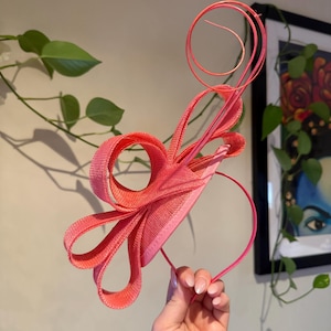 May include: A coral-colored headpiece with a headband base. The headpiece features a circular design with a looped bow and a long, spiraling accent. The accessory is held up against a neutral background with green foliage.