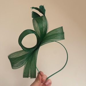 May include: A dark green headband with a bow-like design and a small feather accent. The headband features a looped design and is held by a hand. The accessory is likely for special occasions.