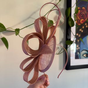 May include: A dusky pink fascinator with a looped design and fine wire detailing. The headpiece has a woven texture and is held by a slender wire stem. This elegant accessory is suitable for special occasions.