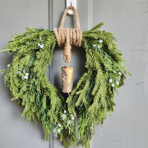May include: A green wreath with faux evergreen branches and small white berries. The wreath is adorned with a rustic, gold-colored bell and a jute rope hanger, perfect for seasonal home decor.