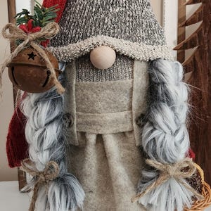 May include: A handcrafted gnome doll with a red knit cap, gray and white knit hat, and long gray braids. The gnome wears a tan felt outfit and brown felt shoes. It holds a bell and a small basket, perfect for holiday decor.