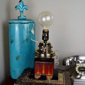 Peut inclure: Une lampe fantaisiste réalisée à partir de matériaux récupérés, avec une ampoule en verre clair, une base dorée et une boîte rouge et dorée. La lampe est posée sur une surface en bois à côté d'un téléphone à cadran argenté.