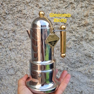 Pode incluir: Uma cafeteira espresso de metal prateado polido com alça e botão da tampa dourados. Uma etiqueta em forma de diamante presa à alça apresenta o texto KONTESSA e 24K. O texto museum italy é visível em amarelo no canto superior direito.