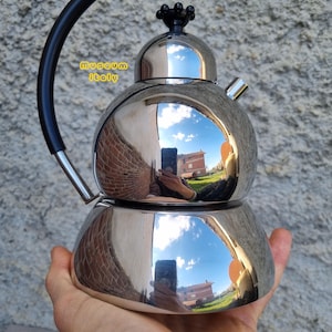 Op de afbeelding: Een gepolijste roestvrijstalen espressomaker met een zwarte handgreep en een decoratieve zwarte knop. Het oppervlak reflecteert de omgeving. De tekst "museum italy" is zichtbaar.