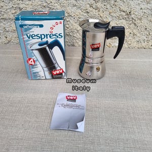 Op de afbeelding: Een roestvrijstalen VEV Vespress espressomaker met een zwarte handgreep en een blauw-witte doos. Op de doos staat "caffettiera" en "Vespress". Er is ook een klein wit papiertje met tekst aanwezig.