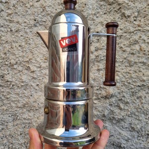 Op de afbeelding: Een roestvrijstalen espressomaker met een houten handvat en bovenkant. De koffiezetter heeft een rood-witte "Vev Vigano" sticker op de voorkant. De koffiezetter wordt in een hand gehouden.