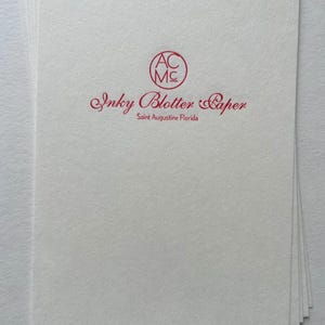 May include: A stack of white Inky Blotter Paper sheets with a red logo and text. The logo is a circle with the letters "AC MC" inside. The text reads "Inky Blotter Paper" and "Saint Augustine Florida".