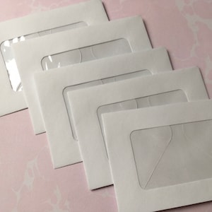 May include: A stack of five white envelopes with clear, rectangular windows. The envelopes are arranged diagonally, with the windows facing the viewer. The background is a light pink, marbled surface.