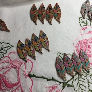 May include: Wooden feather-shaped buttons with colorful patterns and small black buttons. The buttons are arranged on a white surface with pink rose embroidery and green leaves.