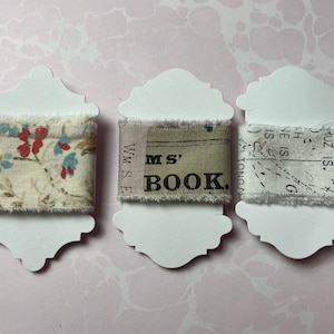 May include: Three spools of decorative fabric ribbon are displayed on white cardstock tags. The ribbons feature floral, text, and map patterns in various colors. The ribbon is frayed on the edges.