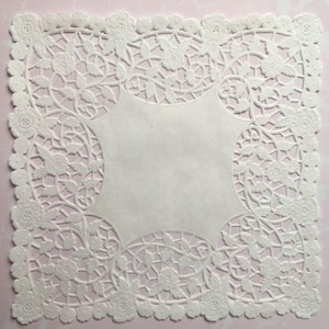 May include: A white paper doily with a floral and vine design. The doily has a scalloped edge and a blank, square center. The intricate cut-out pattern is visible against a pink background.