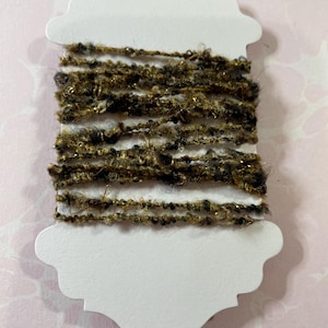 May include: A spool of textured yarn in shades of brown, black, and gold, displayed on a white, decorative tag. The yarn has a fuzzy, uneven texture, with metallic gold threads interwoven throughout. The tag has a scalloped edge and a small loop at the top.