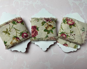 May include: Three rolls of floral burlap ribbon. The ribbon features a beige background with a repeating pattern of pink and yellow roses and green leaves. The edges are frayed, and the rolls are mounted on white cardstock.