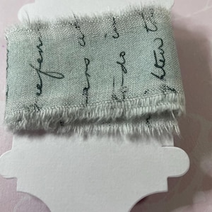 May include: A light sage green fabric ribbon with frayed edges wrapped around a white cardstock spool. The ribbon features a repeating black cursive script pattern printed across the surface, creating a textured and detailed appearance.